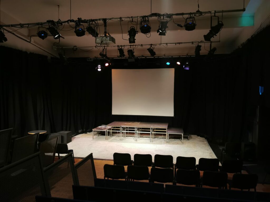 An image of our stage as taken from the back row of the seats. A projector screen hangs down and some stage blocks are erected