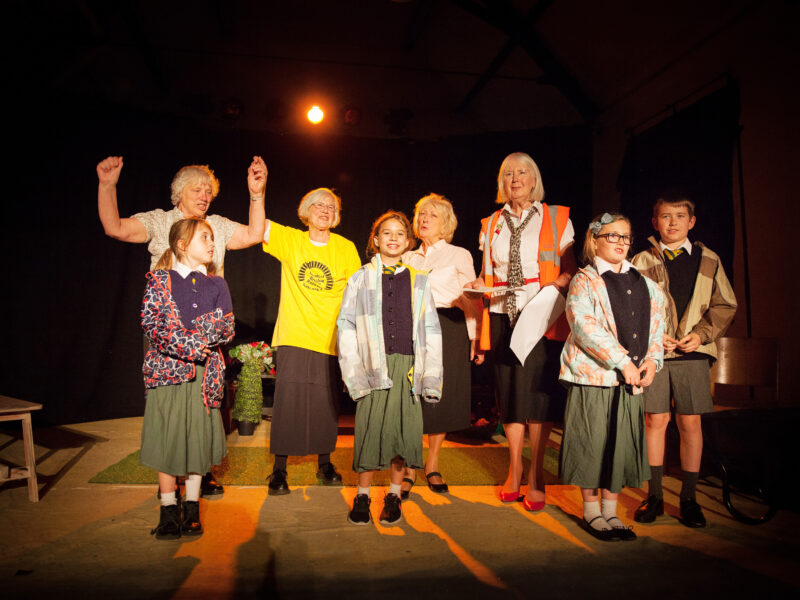 An intergenerational group of actors stand on stage