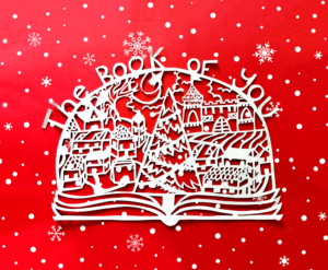 A papercut of a christmas village, surrounding a christmas tree, inside a snow globe mounted on a book