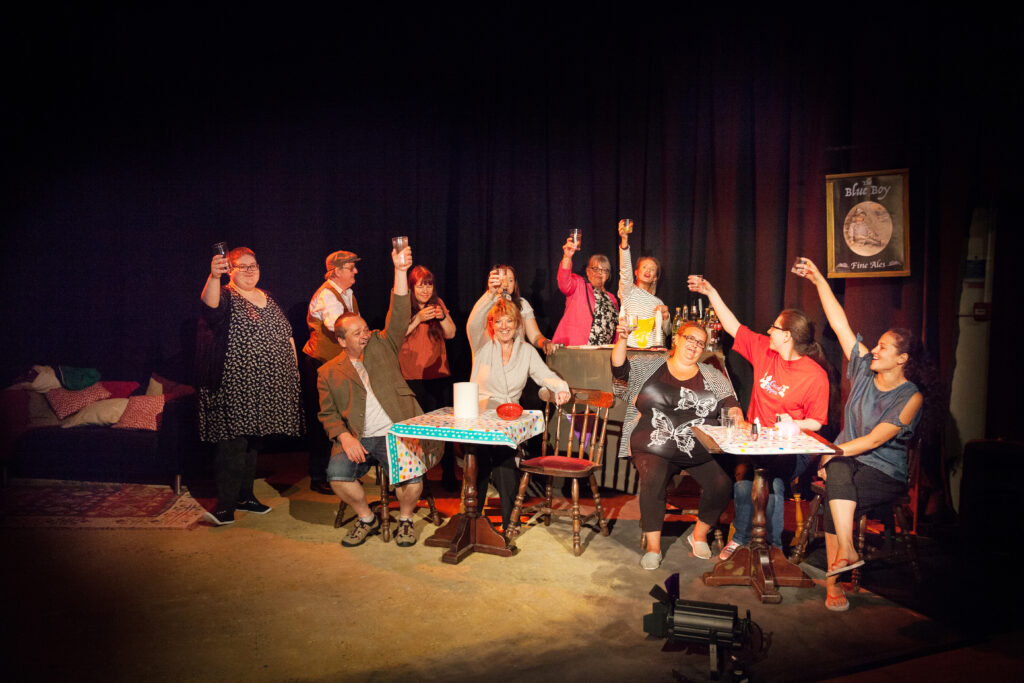 A stage with a pub in it, numerous actors on stage holding up drinks in a celebratory fashion