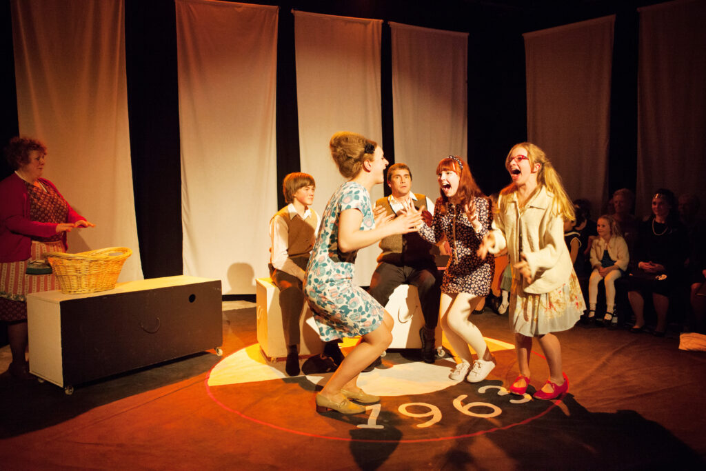A group of people stood on stage dressed as though it is the sixties, jumping and screaming with excitement