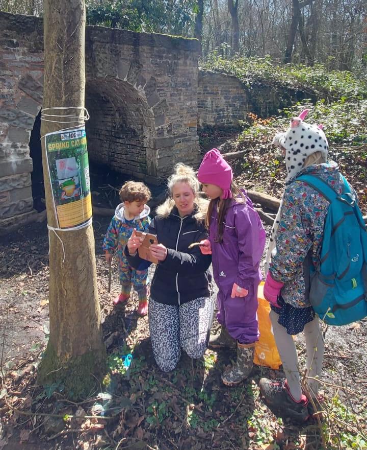 One adult crouches to help read a sign to three children in a woodland area