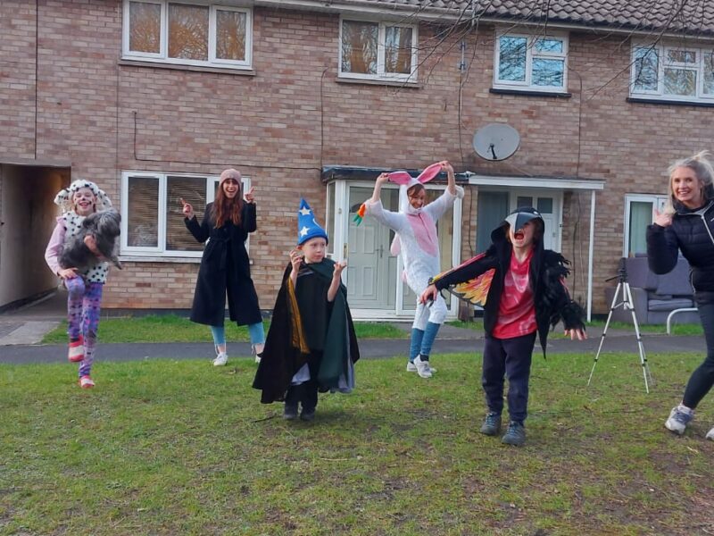 Seven people, three of whom are adults and four children, stand in differing costumes in front of a row of houses