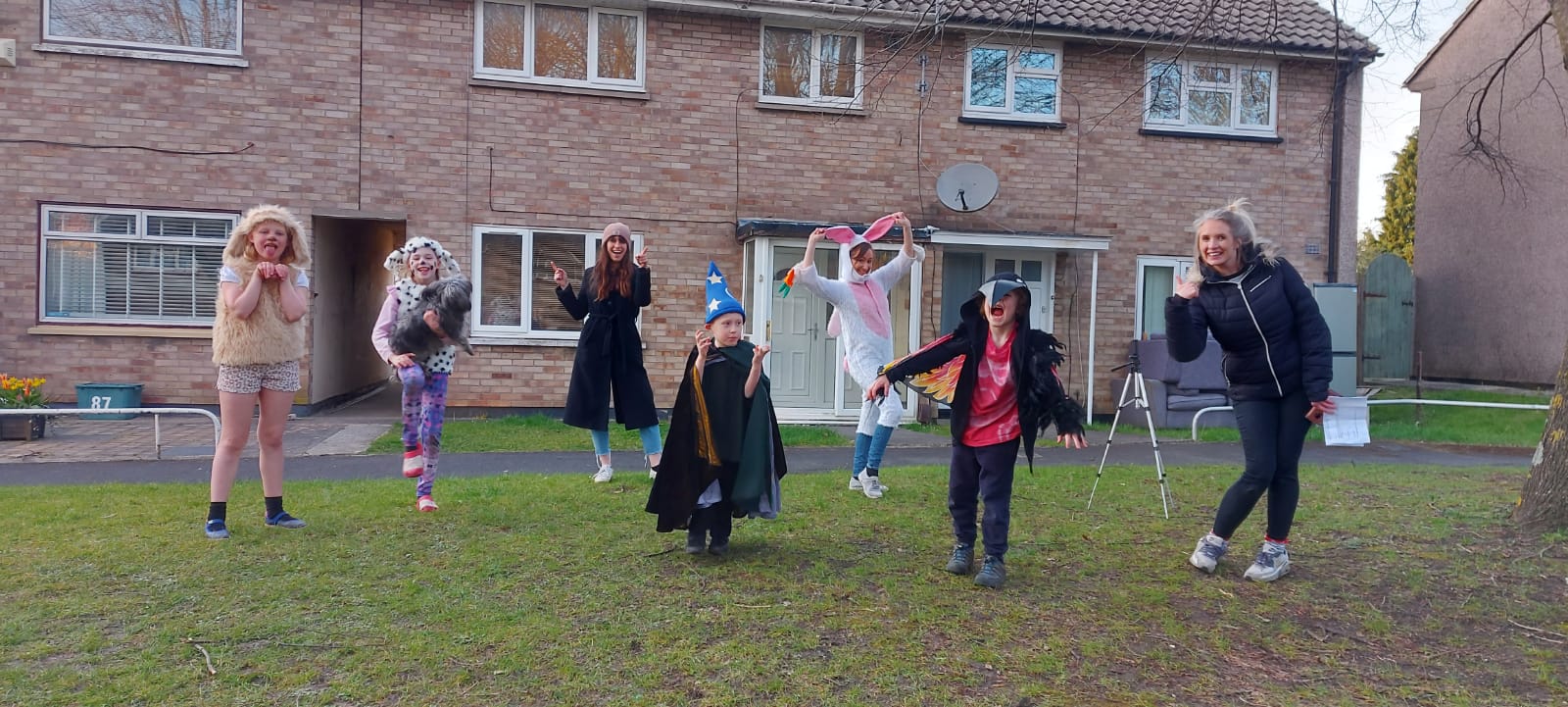 Seven people, three of whom are adults and four children, stand in differing costumes in front of a row of houses