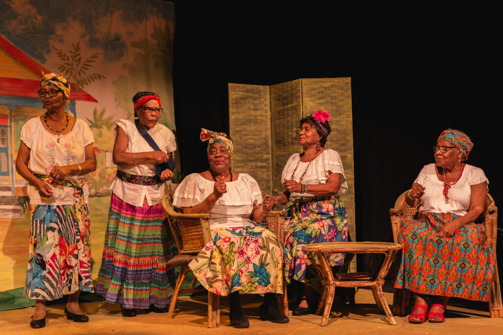 Five actors in colourful skirts sit on a stage with bamboo furniture