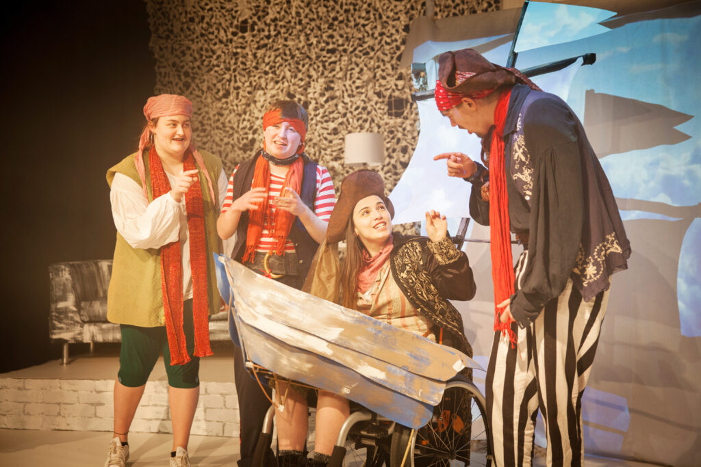Four actors dressed as pirates, and one using a wheelchair dressed as a boat, stand on stage