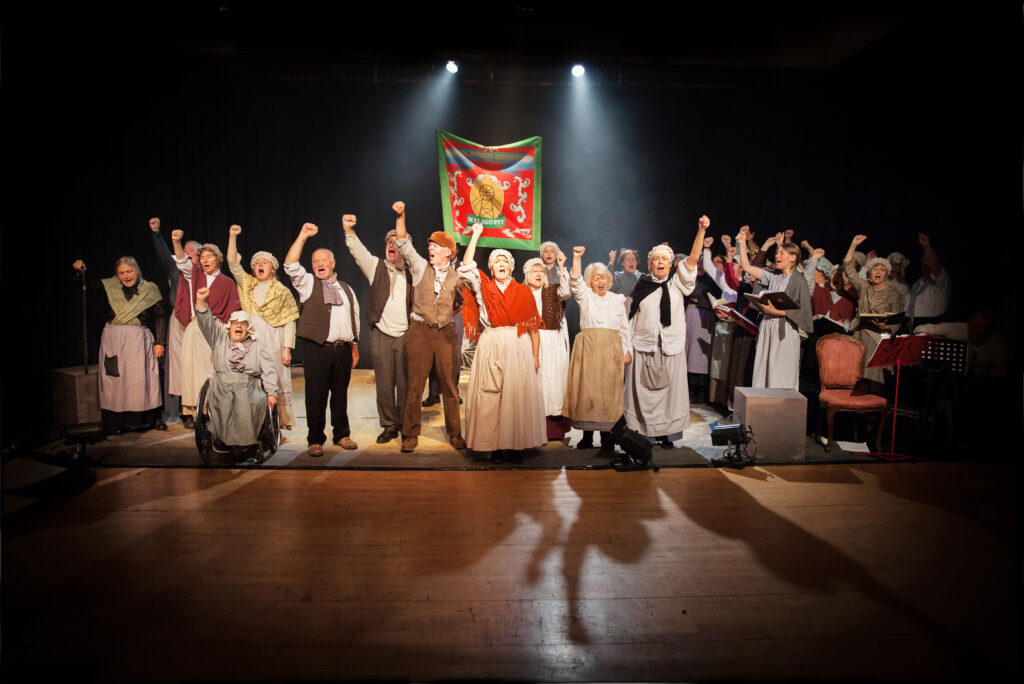 A group of actors dressed as Victorians, stand with their fists in the air in an act of triumph