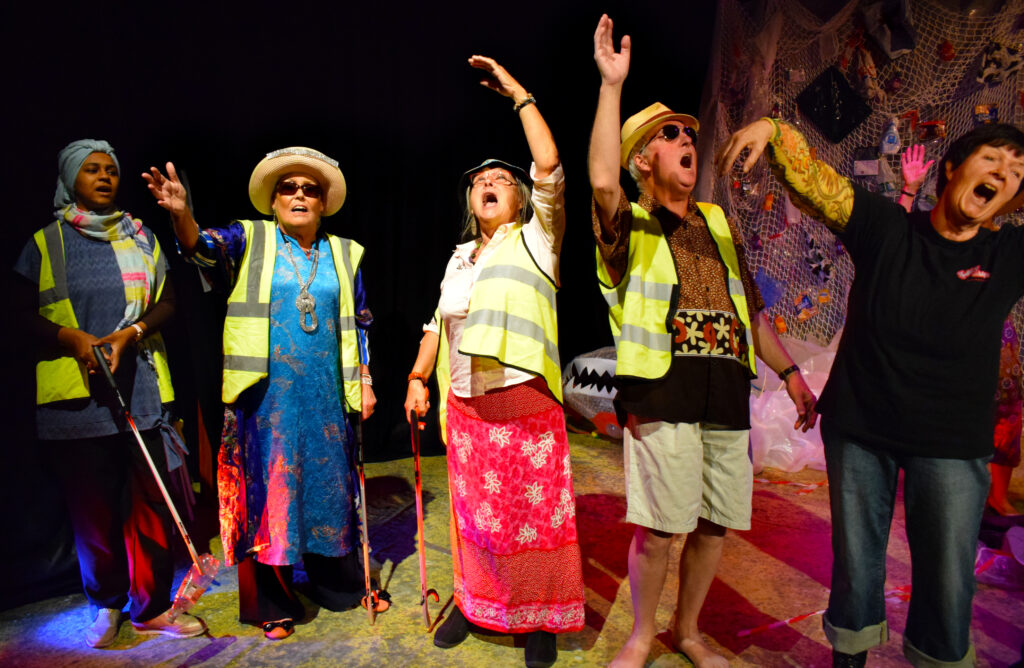 Five actors in high vis jackets, holding litter pickers stand on stage with their hands in the air and an exasperated look on their faces