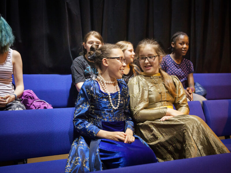 Two young actors take a break, sitting in the theatre seats chatting whilst wearing mediaeval looking gowns