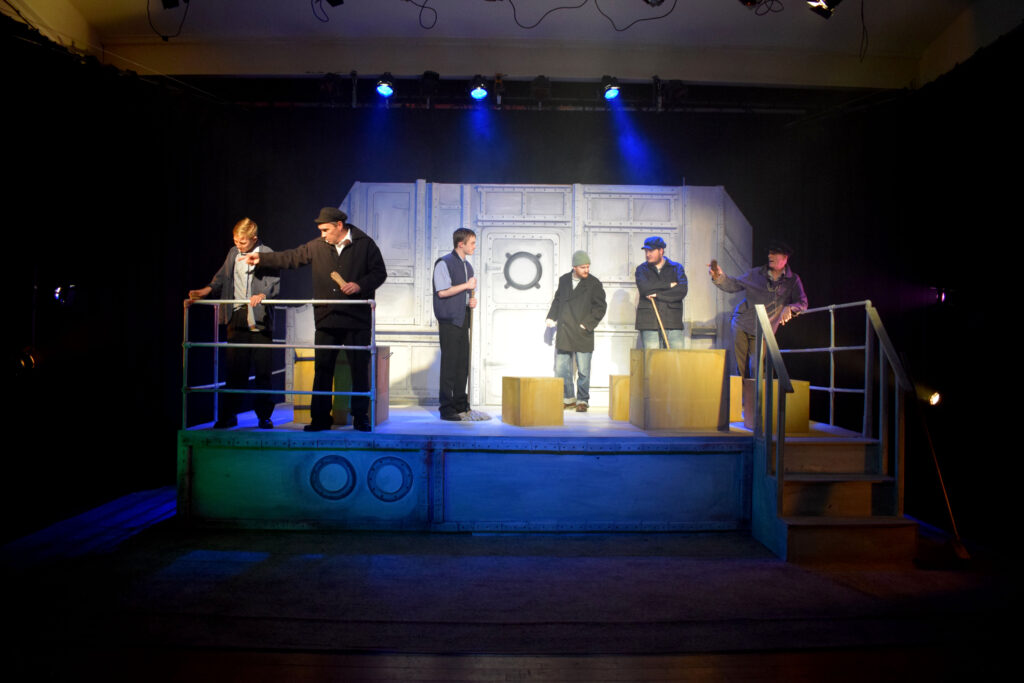 Six actors stand on a stage decorated to look like the top deck of a boat