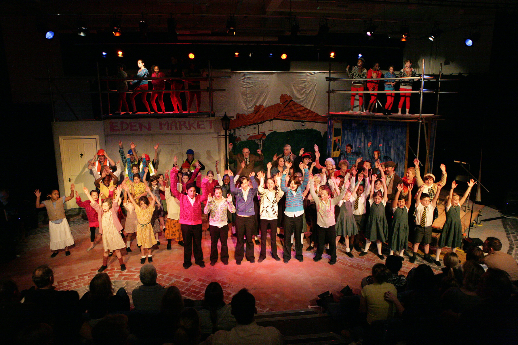 A large cast stand in a big stage set, all raising their hands in the air in triumph