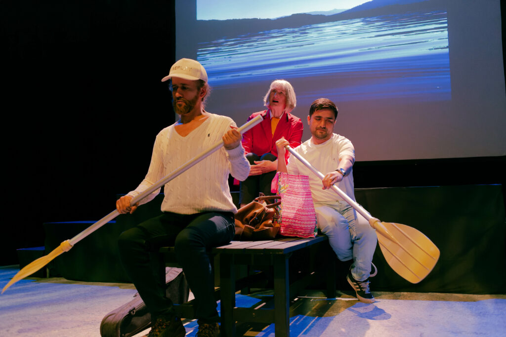 Three actors on stage, sat on a bench and using canoe paddles to suggest they are boating