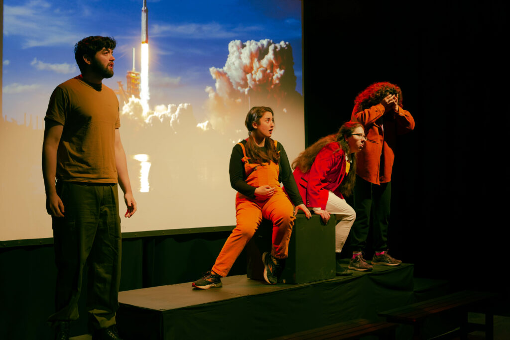 Four actors on stage in front of a projection of a rocket launch, looking shocked