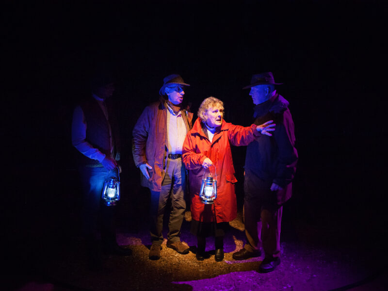Four actors stand on a dark stage, illuminated by two hand-held lanterns. All actors are wearing outdoor wear and looking worried