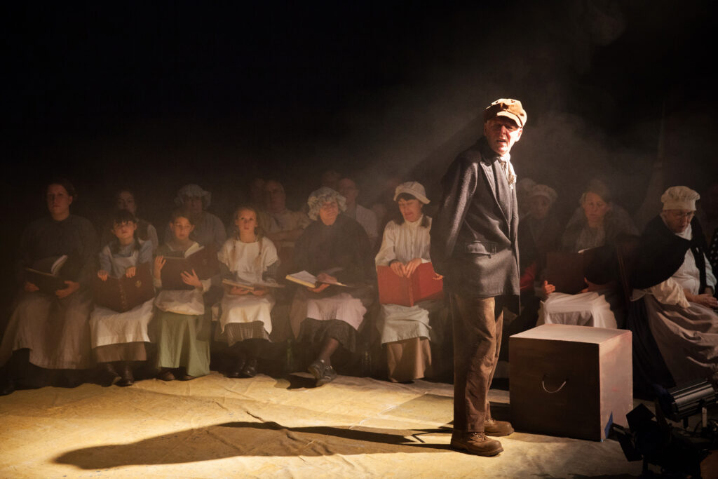 An actor dressed as an 1800s miner, stood on stage. The front row is a choir also dressed in 1800s clothes.
