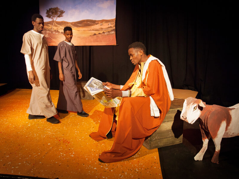 Three actors on stage, one is reading the newspaper. All are wearing loose fitting robes. A model of a goat stands to the side and there is a projection of a desert on the back wall
