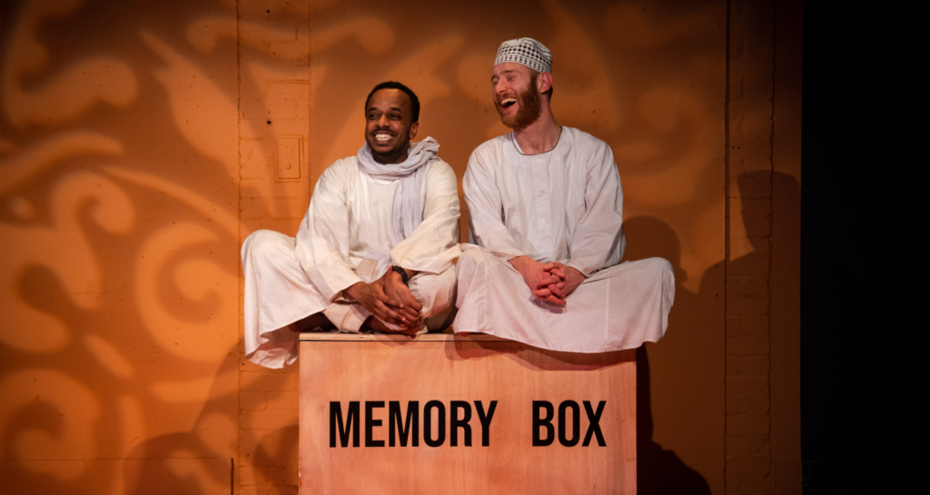 Two men sit on top of a wooden box labelled 'Memory Box', smiling. They are both wearing white robes