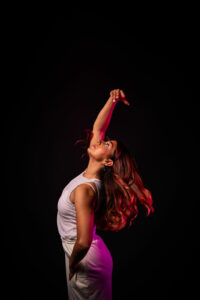 A dancer stands up, with one arm in the air, her back arched and she is looking directly upright.