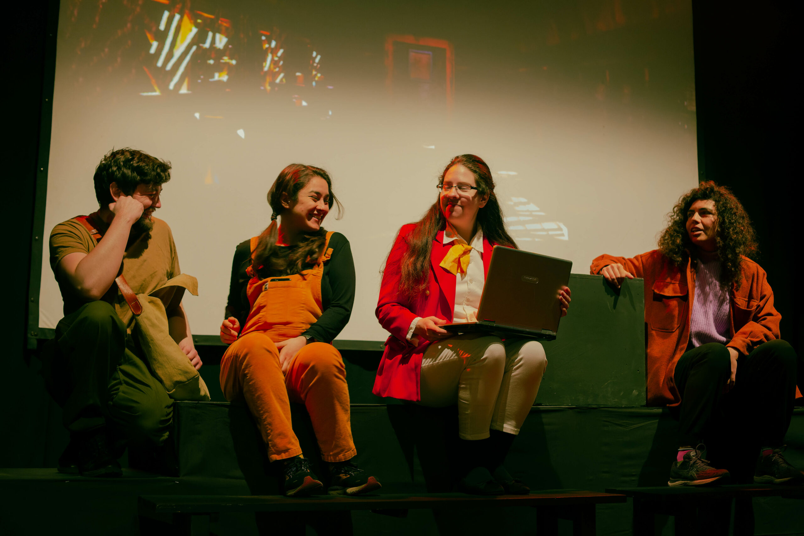 Four actors sit on stage, laughing and smiling with one another. One actor is using a laptop