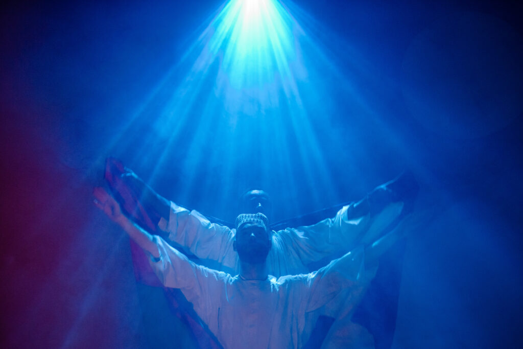 Mohand and Peter stand bathed in a blue spotlight, with their arms outstretched. Their faces have a neutral, strong expression. 