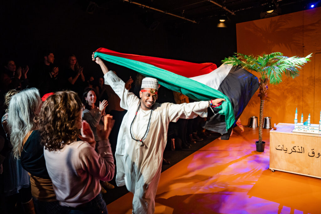 Mohand runs around the stage, smiling holding the Sudanese flag aloft, whilst the audience stand and applaud. 