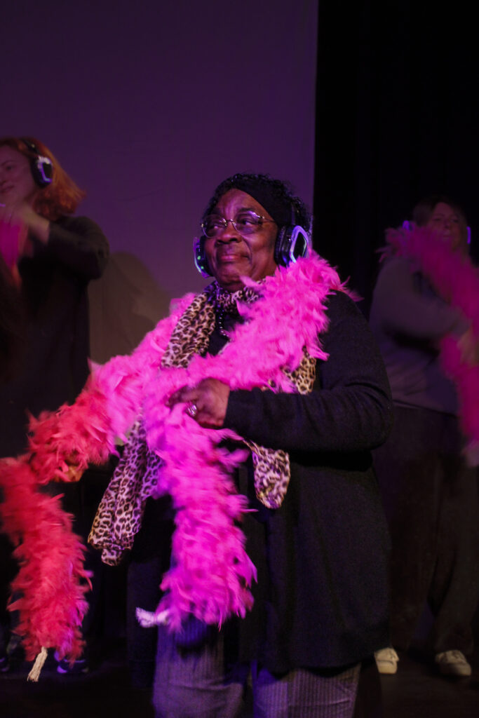 An actor wearing over-ear headphones dances whilst wearing a pink feather boa