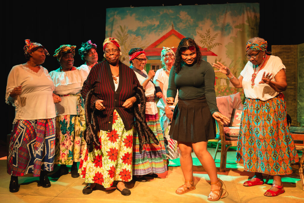 The actors, wearing colourful ankara skirts, dance together. One actor is wearing a cardigan designed to make them look like a spider