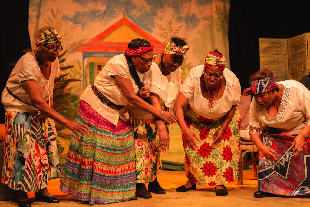 The actors, wearing colourful ankara skirts, mime warming their hands on a fire