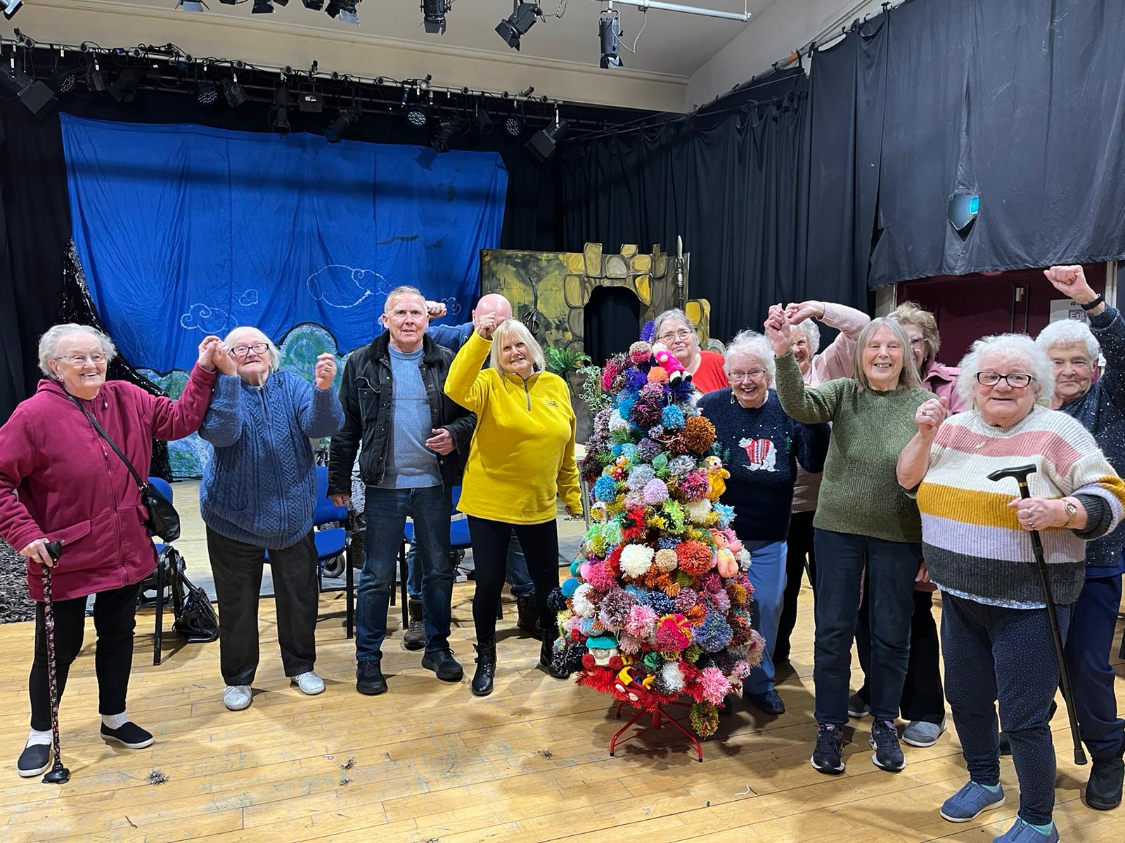 Eleven members of our over 55s group stand around a Christmas tree made of brightly coloured pompoms, cheering.