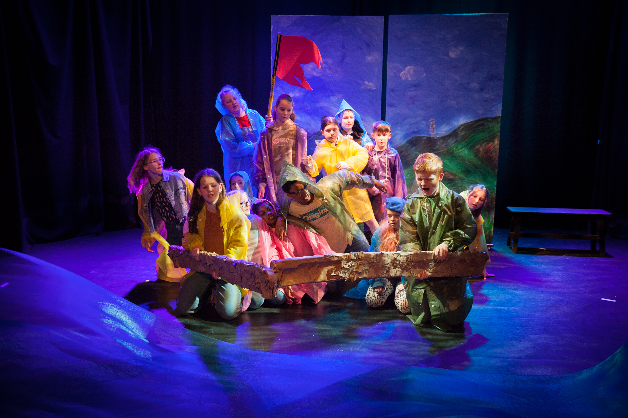 A large group of young actors in raincoats shelter from floods in a home made raft